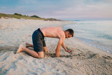 Young topless barefoot man in shorts digging hole by hands on beach beyond sea in summer evening. Funny boy playing on beach with sand on desolate cost near ocean ike child. Vacation leisure and hobby