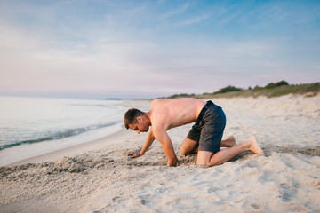 Fototapeta na wymiar Young topless barefoot man in shorts digging hole by hands on beach beyond sea in summer evening. Funny boy playing on beach with sand on desolate cost near ocean ike child. Vacation leisure and hobby