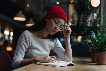 Portrait of tired university student studying, learning language, exam preparation, reading, taking notes sitting in library. Asian woman wearing hipster hat working project, busy, serious face. 