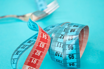 Tape measure around a fork as concept for diet. Fork are wrapped in blue measuring tape on blue background.