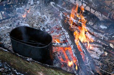Black hiking tourist pot, which is brewed mulled, stands on the coals of the fire. Picnic. Warm...