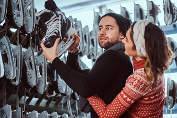 Young couple wearing warm clothes standing near rack with many pairs of skates, choosing his size, preparing to skate on the ice arena.