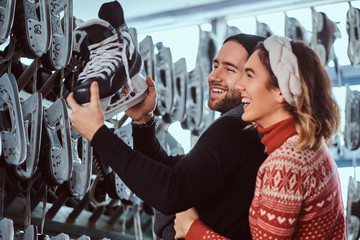 Cheerful young couple wearing warm clothes standing near rack with many pairs of skates, laughing...