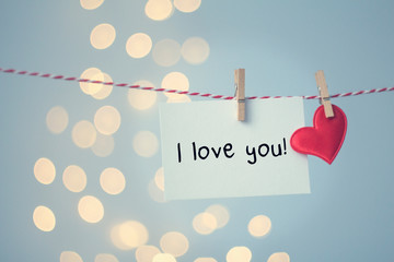 A note saying "I love you!" and a red heart hang on a string with clothespins against a bokeh background. Concept of: Expressing affection