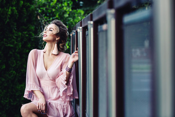 attractive young girl in a short pink dress posing near a steel fence on the background of green foliage.