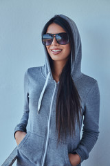 A beautiful girl wearing a gray hoodie and sunglasses holds hands in pockets, smiling and looking at a camera in front of a wall outdoors