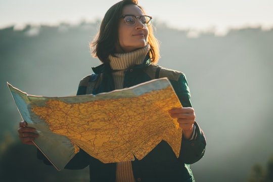 Happy smiling traveller girl holding big map and preparing a route for her trip around the mountains and looking around, focus on the face, blurred trees and forest in the background