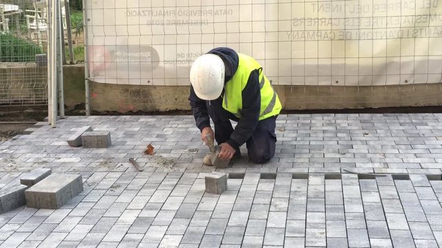 Public Ajuntament de Barcelona worker with hammer, helmet and yellow jacket hammering a square block of concrete on under construction pedestrian street or sidewalk