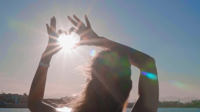The girl make heart with her hands over sea background. Silhouette hand in heart shape with sunset inside. Vacation concept.