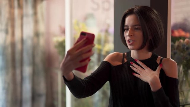Very beautiful hot sexy girl with short black hair in black shoulderless sweater standing near window in flower shop, doing videocall, showing flowers, talking and gesturing. Close-up concept