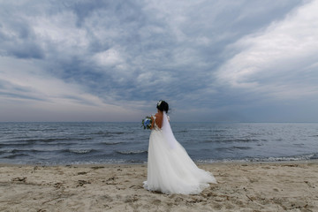 Fototapeta na wymiar woman in long white dress with a blue bouquet of flowers outdoor on sea beach. Stylish bride in Dress with naked back ang long veil on the river bank. at summer