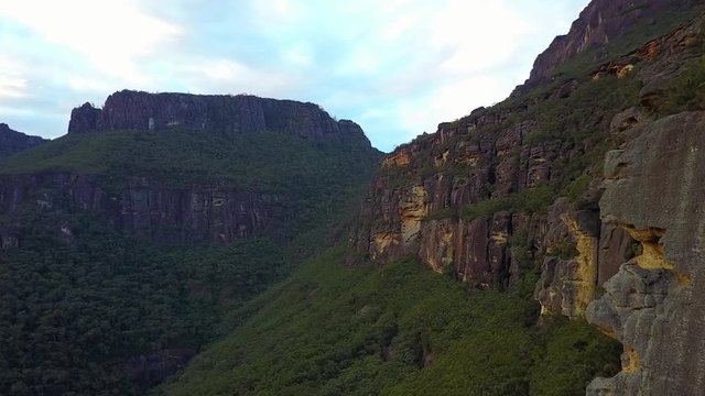 Drone flying alongside orange and black cliff face with large rock cliffs in the background.