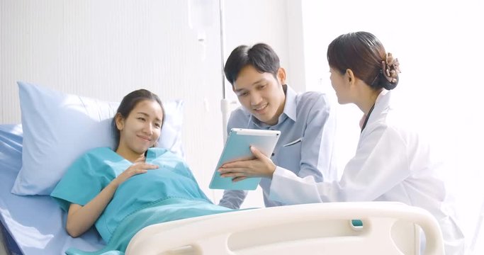 Lovely asian couple excited parents-to-be look at sonogram pictures on digital tablet in hospital room.