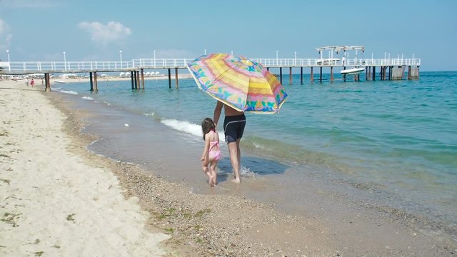 A man with a child walk on the beach. A father and his little daughter walk under an umbrella along the seashore.