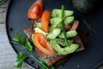 Healthy avocado sandwich on dark rye toast bread made with fresh sliced avocado, seeds and tomatoes on rustic wooden background.