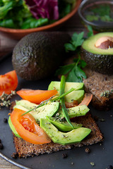 Healthy avocado sandwich on dark rye toast bread made with fresh sliced avocado, seeds and tomatoes on rustic wooden background.