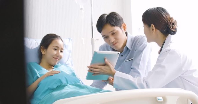 Lovely asian couple excited parents-to-be look at sonogram pictures on digital tablet in hospital room.