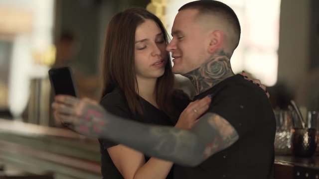 Couple is in caf&radic;&copy;. They are happy together and making selfies. The guy is biting the girl for a cheek and than they are kissing on camera. Both of them  have a lot of tattoos on neck and hands.
