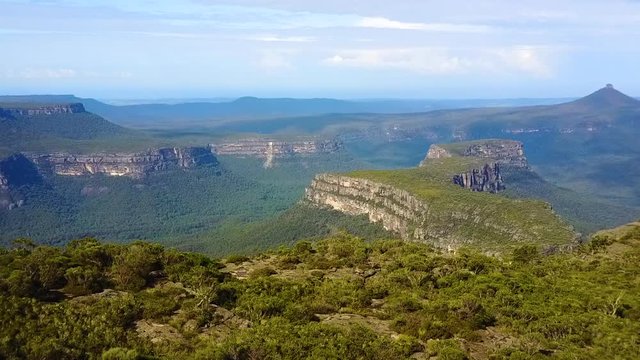 Drone flying over tree lined cliff edge revealing deep valley, mountains and rocky escarpments. Location NSW, Australia.