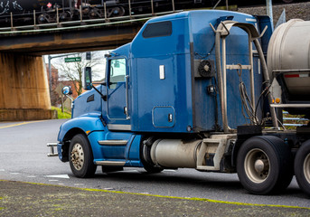 Blue American bonnet rig semi truck with tank semi trailer for transportation of liquids and chemicals running on the road under the bridge