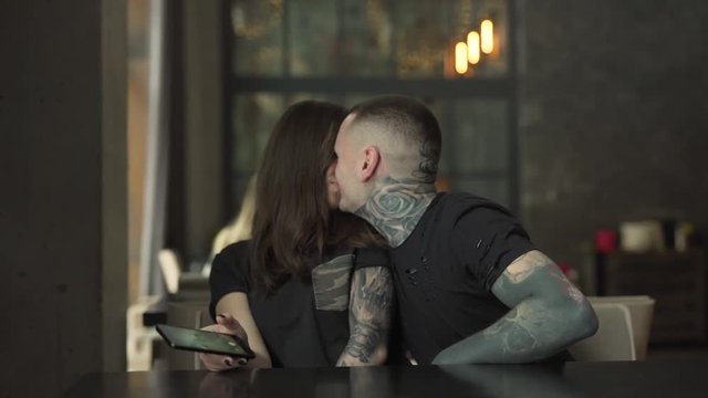 Couple is in caf&radic;&copy;. They are happy together and making selfies.They are kissing on camera. Both of them  have a lot of tattoos on neck and hands.