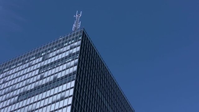 An antenna high on top of a modern skyscraper. Bright blue clear sky. Tall glass building reflects the daytime sun and sky giving a blue appearance.