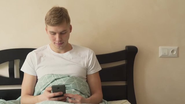 Handsome blond man lying in bed, surfing/scrolling/using his phone. His girlfriend/woman coming up to him with a cup of coffee, hugging and kissing him. They smiling, satisfied. Lovers/couple concept