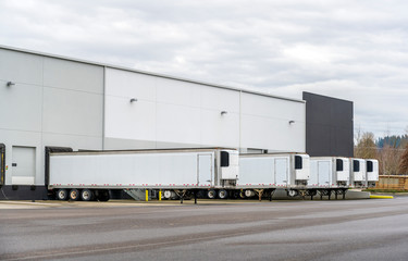 Long refrigerator semi trailers stand in row in warehouse docks with gates and loading cargo