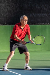 Focused Chinese elderly man ready with racquet raised and serious expression during a game of tennis.