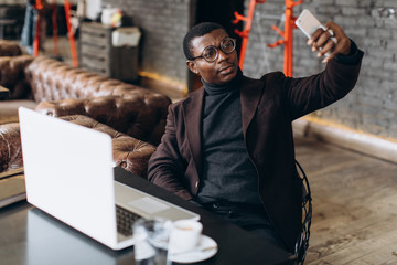 Portrait of happy african businessman using phone and make selfie while working on laptop in a restaurant.