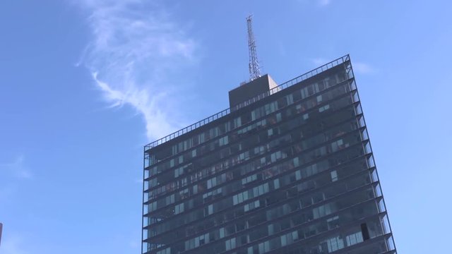 Thin white clouds blow past the top of the Kista Science Tower. Sharp angles and walls of glass in the design of this modern skyscraper towering above other buildings in the area.
