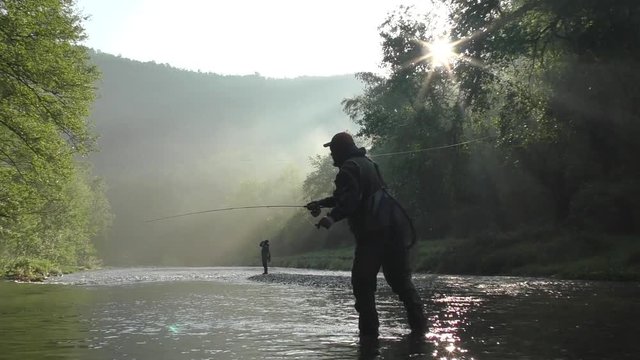 two fly fishermen silhouette catch fish with rods on small river in early summer morning at rays of rising sun