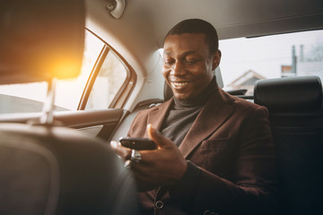 Young smiling african man using smartphone while sitting on backseat in car. Concept of happy...
