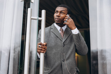 Handsome young african american businessman in classic grey suit holding a smartphone and smiling while leaving the office building