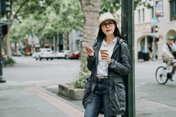 Obraz premium Beautiful girl walking around the city with a cup of coffee smiling. office lady relying on street lamp using mobile phone waiting for friends hold hot tea after work. man riding bike on background.