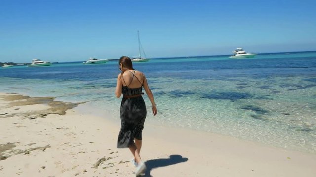 Brunette walks along white beach with blue water and yachts in background SLOMO