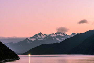 Breathtaking pink skies at sunset with snow capped mountains and the light from a vessel sailing in the distance in Skagway, Alaska