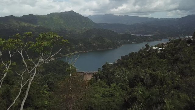 AERIAL: pushing past a skinny tree to reveal a dam and lake between the mountain tops of Utuado, Puerto Rico.