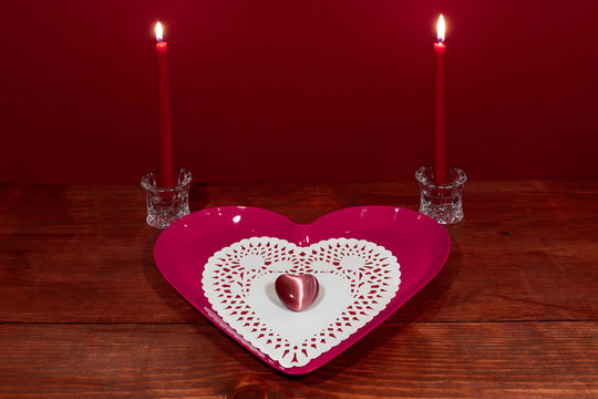 Pink heart shapped plate with dollie and gemstone, two red candles in crystal holoders on wooden table.