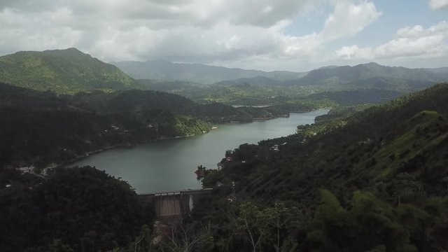 AERIAL: pushing over some shrubbery to reveal the dam and lake within the valley between the mountain tops of Utuado, Puerto Rico.