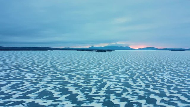 Aerial SLIDE over a frozen lake with snowy patterns in the ice and sunrise color poking through the mountain range on the horizon