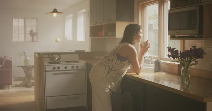A woman stands looking out the window of her kitchen into the sun holding a mug.  A vase of purple flowers sits on the countertop next to her.