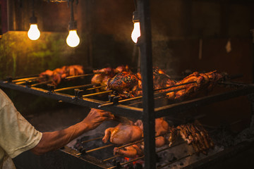 outdoor street chicken barbecue grill arm and hand brush oiling the grilling meat food broiler spit roasted over smoking living coals in a simple electric illumination, the Philippine lechon Manok