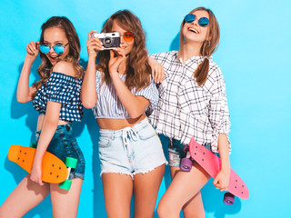 Three sexy beautiful stylish smiling girls with colorful penny skateboards.Women in summer hipster checkered shirt clothes posing near blue wall. Models taking pictures on retro camera