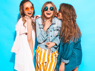 Three young beautiful smiling hipster girls in trendy summer casual clothes. Sexy women share secrets, gossip.Isolated on blue. Surprised face emotions