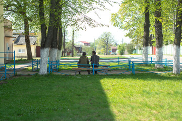 two men sit on a bench in the street in summer in a green park near houses