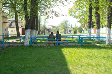 Fototapeta premium two men sit on a bench in the street in summer in a green park near houses