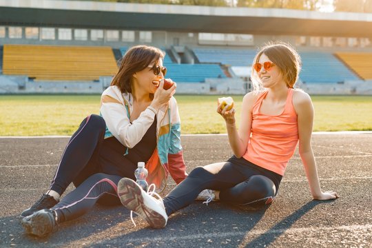 Healthy lifestyle and healthy food concept. Smiling fitness mother and teen daughter together eating apple sitting on stadium after training on sunny spring day
