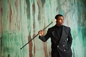 Stylish african american gentleman in elegant black jacket, holding retro walking stick as cane flask or tippling cane with golden diamond ball handle. Rich fashionable afro man against green wall.