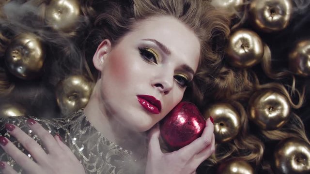 Girl in luxurious golden dress with voluminous beautiful hair lies among the golden apples on black cloth. Smoke. Snow White. Beautiful model with  professional make-up. Concept for advertising.